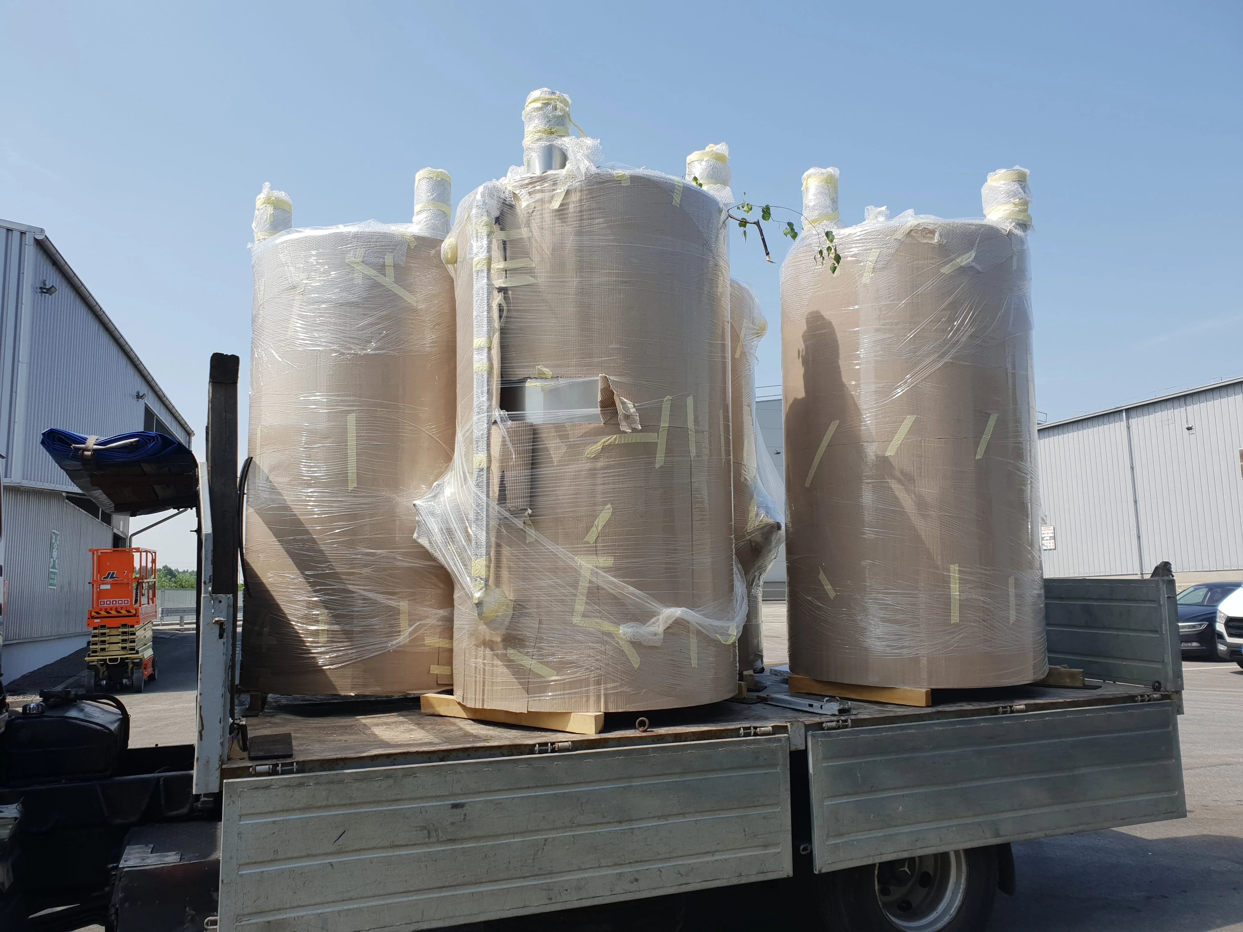 Trzy duże cylindryczne pojemniki owinięte folią na platformie ciężarówki na zewnątrz przy magazynie.