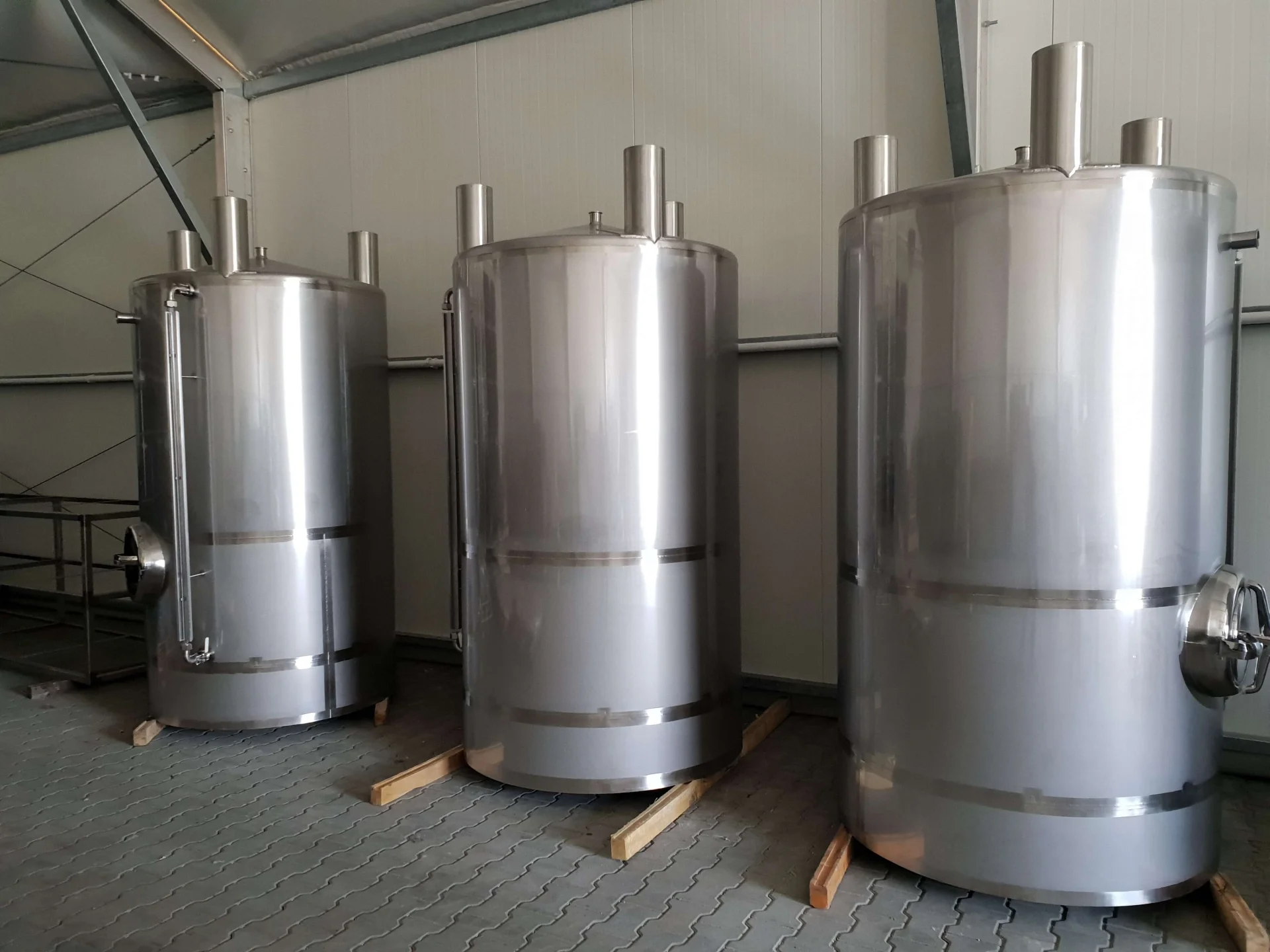 Trzy duże, błyszczące stalowe zbiorniki przemysłowe stojące na drewnianych belkach w magazynie.