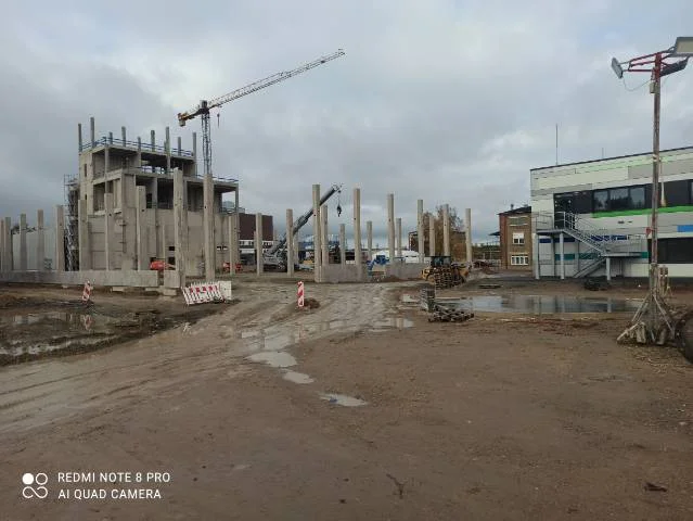 Plac budowy z widocznymi słupami betonowymi, żurawiem i częściowo wybudowanym budynkiem pod pochmurnym niebem.