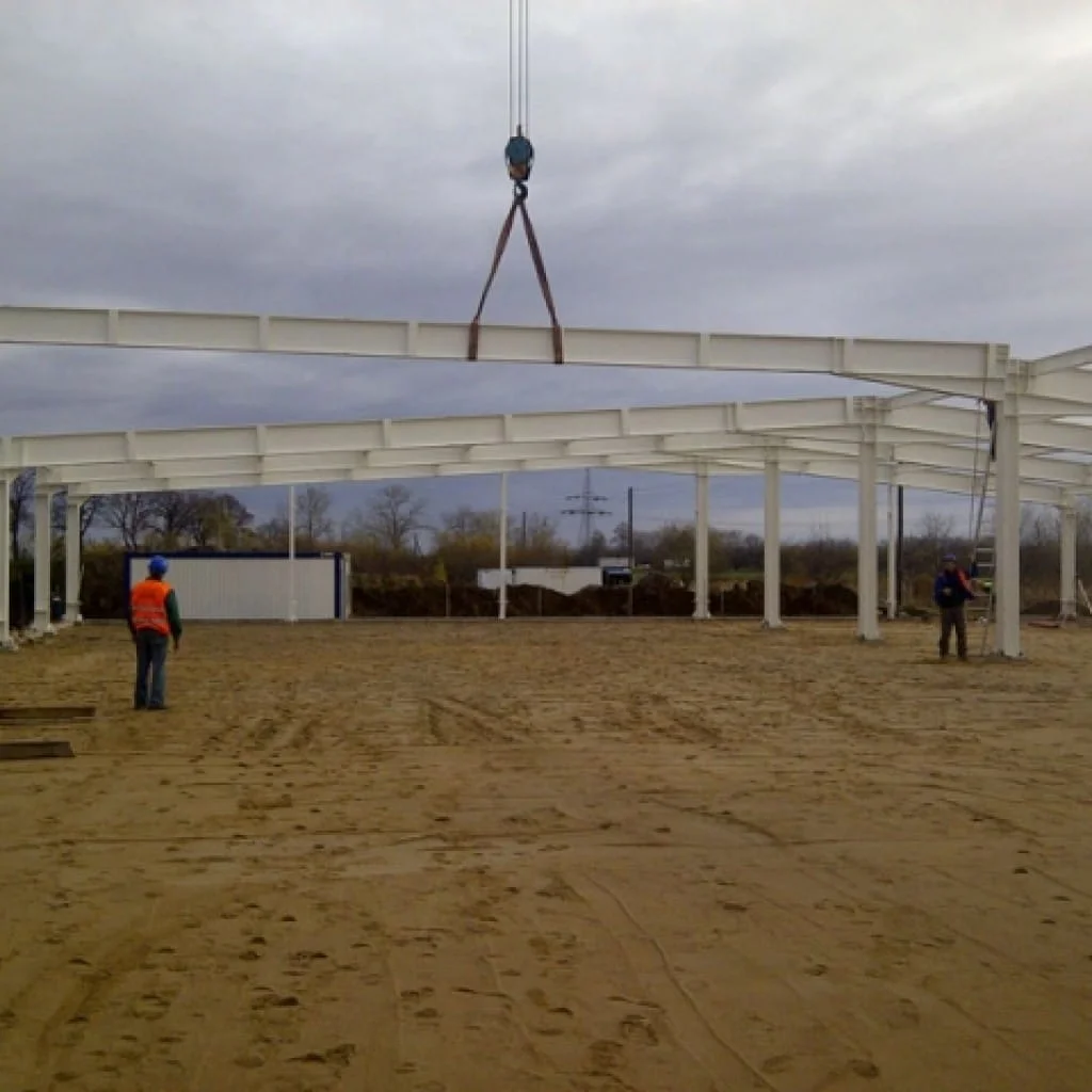 Plac budowy z rusztowaniem i dwoma pracownikami nadzorującymi podnoszenie metalowej belki.