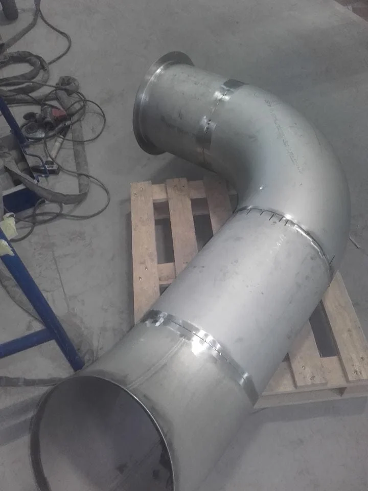 Metalowa rura wentylacyjna zagięta pod kątem 90 stopni ustawiona na drewnianej palecie w hali przemysłowej.