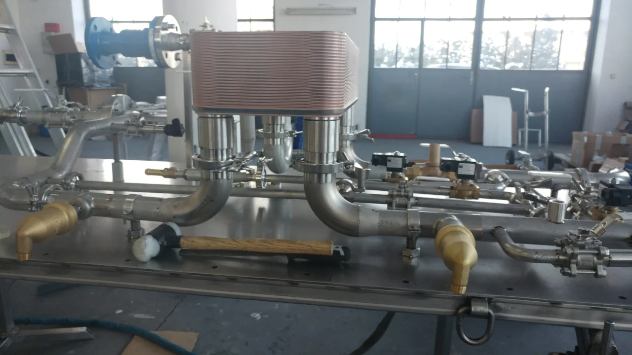 Metalowa aparatura przemysłowa z rurami, zaworami i miedzianym wymiennikiem ciepła na stole roboczym w warsztacie.