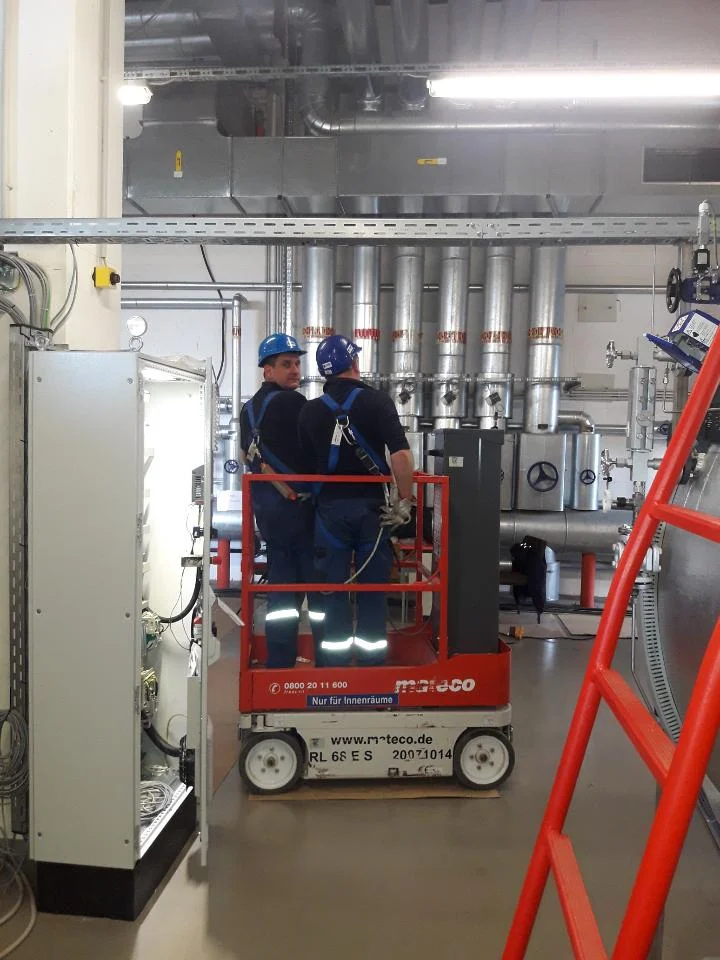 Dwóch pracowników w kaskach i odzieży roboczej stoi na podnośniku w hali przemysłowej z rurami i instalacjami.