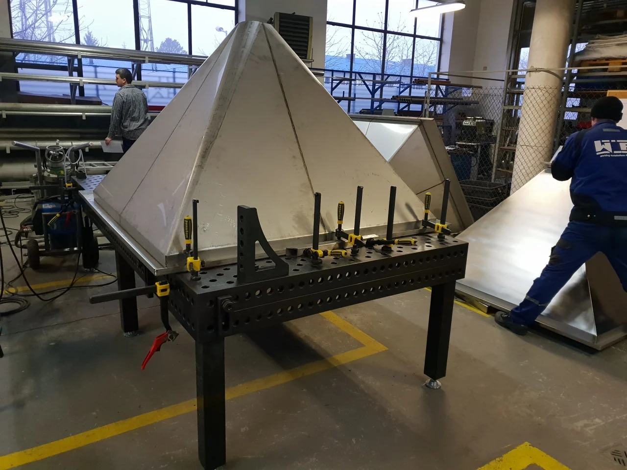 Hala produkcyjna z metalowym stołem roboczym trzymającym dużą metalową piramidę mocowaną za pomocą zacisków.