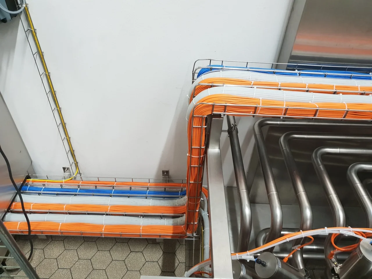 Zorganizowane kable pomarańczowe, niebieskie i białe ułożone w metalowej kratownicy przy ścianie i nad metalowymi rurami.