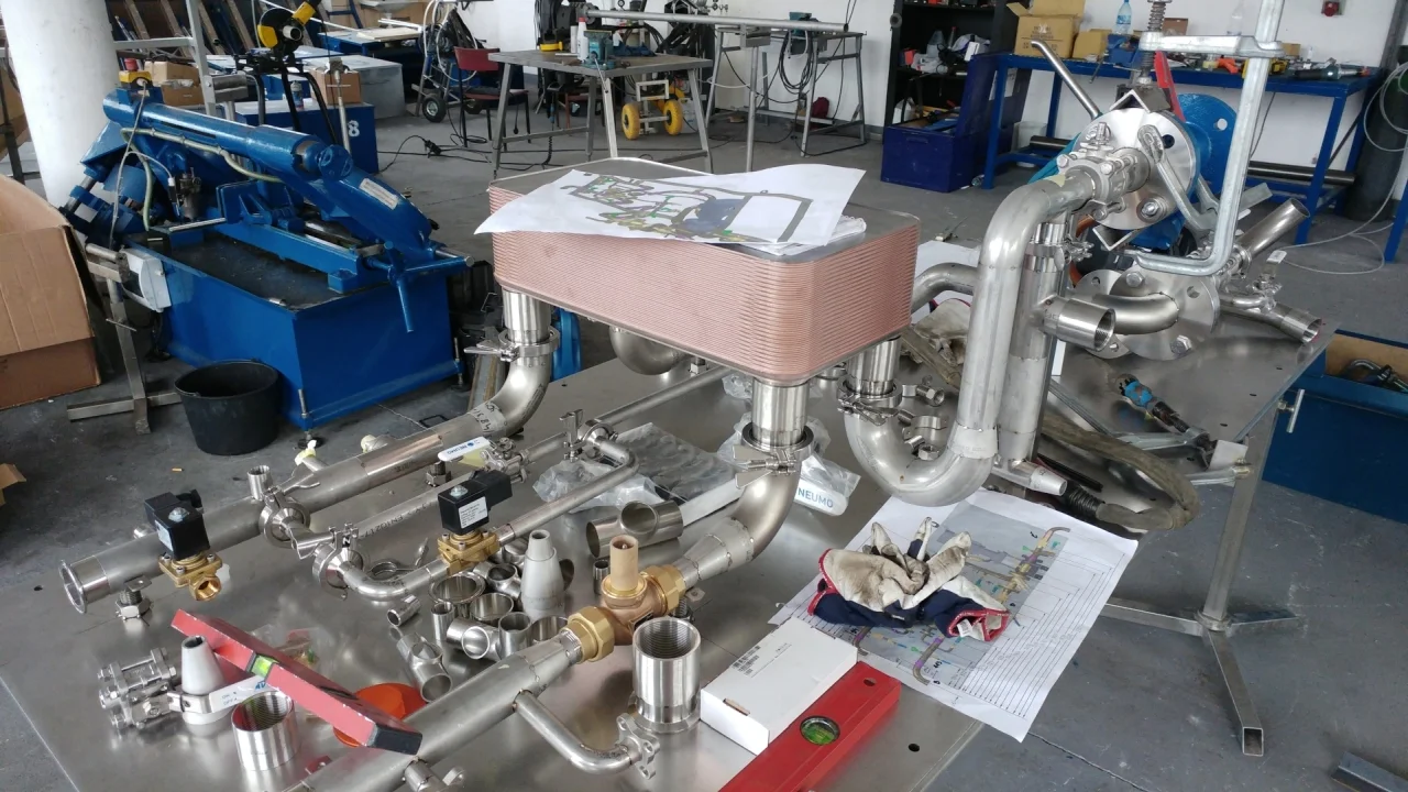 Zespół rur i złączy ze stali nierdzewnej na stole warsztatowym z dokumentacją techniczną i rękawicami roboczymi.