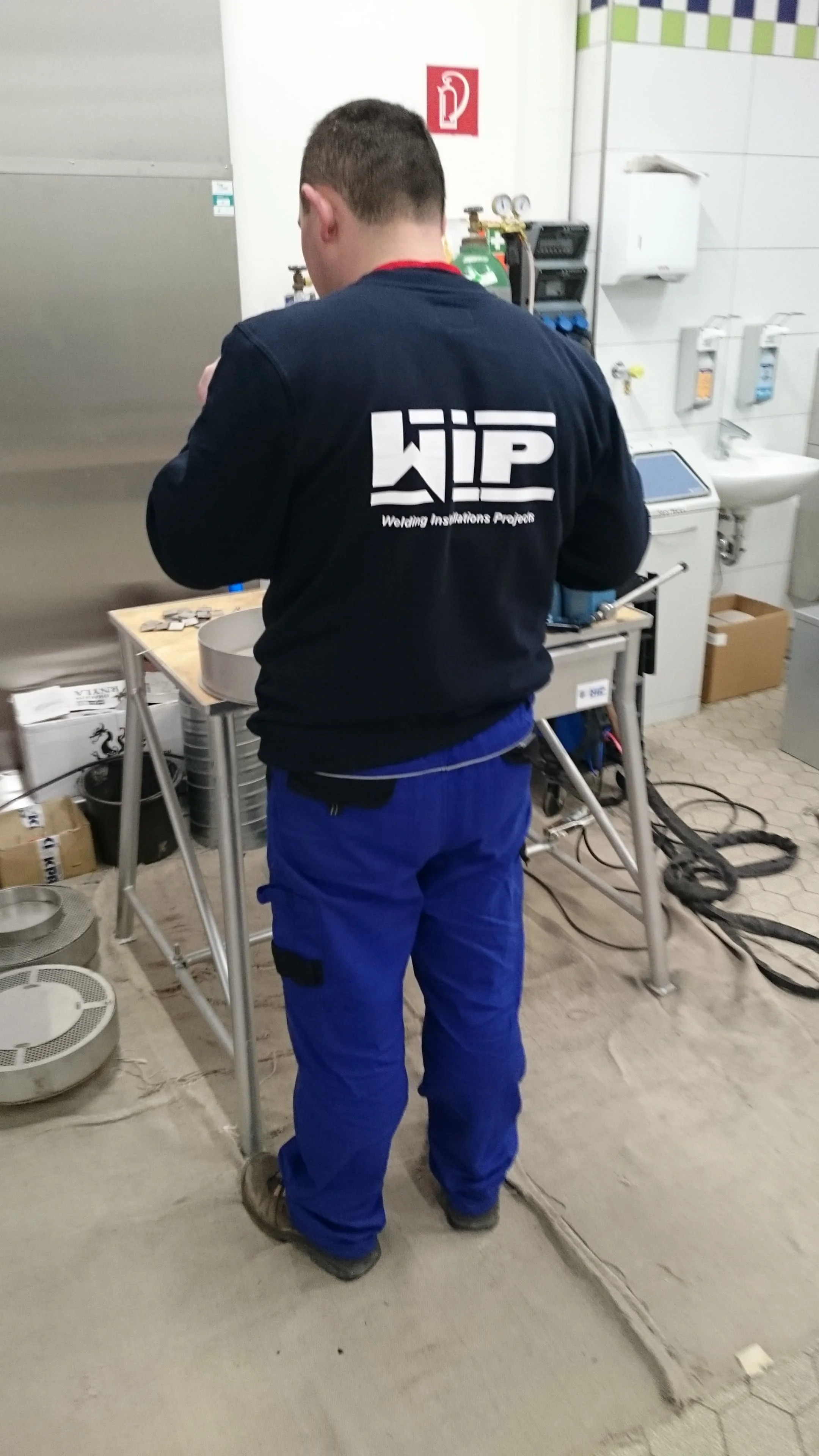 Mężczyzna w niebieskich spodniach i granatowej bluzie z logo WIP stoi przy stole warsztatowym z narzędziami.