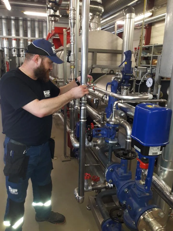 Mężczyzna z brodą i czapką pracuje przy systemie rur i zaworów w przemysłowym zakładzie.