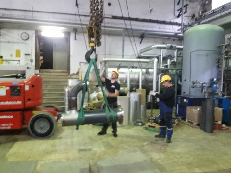 Dwóch pracowników w kaskach operuje dźwigiem przemysłowym w hali produkcyjnej, podnosząc duży metalowy cylinder.