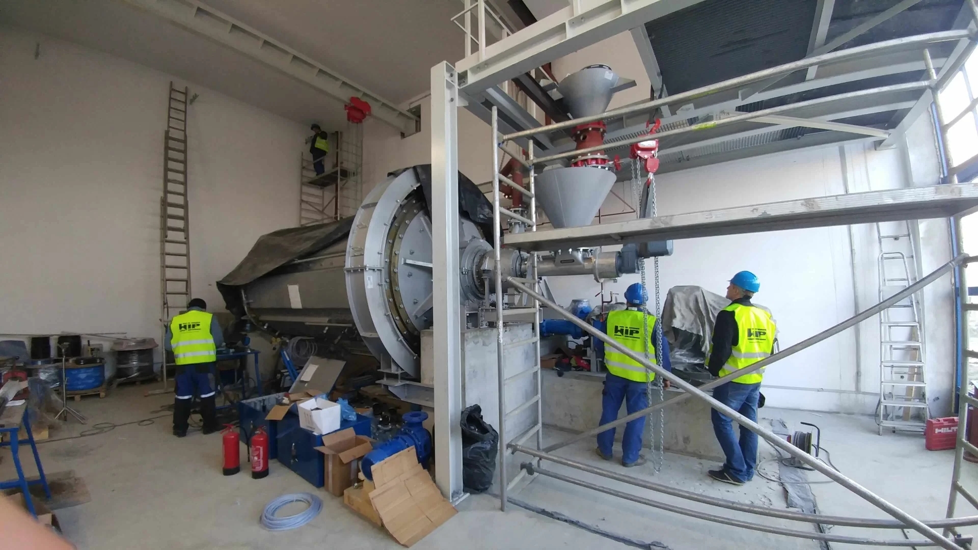 Czterech pracowników w kamizelkach odblaskowych i kaskach pracuje nad dużą maszyną przemysłową w hali produkcyjnej.