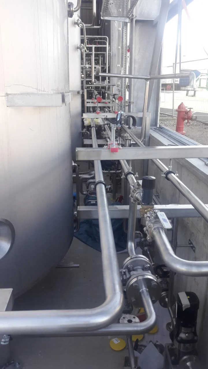 Wnętrze zakładu z trzema dużymi stalowymi zbiornikami przemysłowymi po obu stronach wąskiej platformy z poręczami.