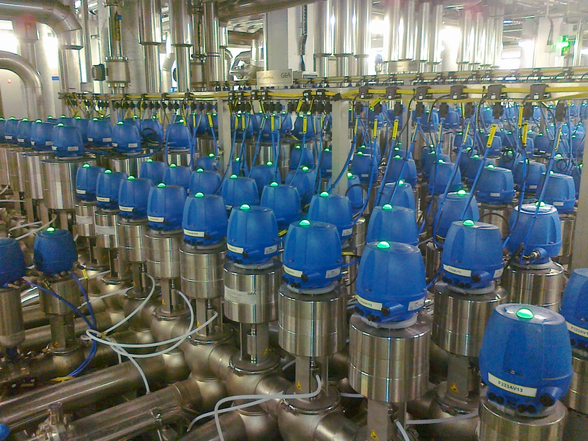 Rząd zautomatyzowanych zaworów z niebieskimi pokrywami i zielonymi wskaźnikami na stalowych rurach w systemie przemysłowym.