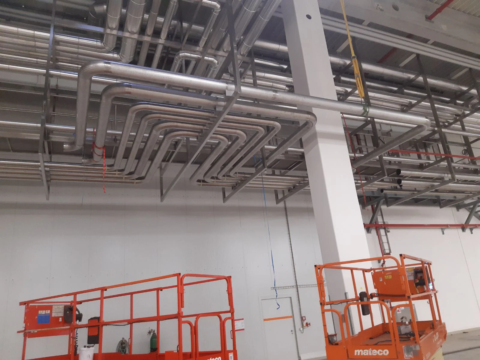 System rur stalowych zamocowanych na suficie w hali przemysłowej z dwoma pomarańczowymi podnośnikami nożycowymi na dole.