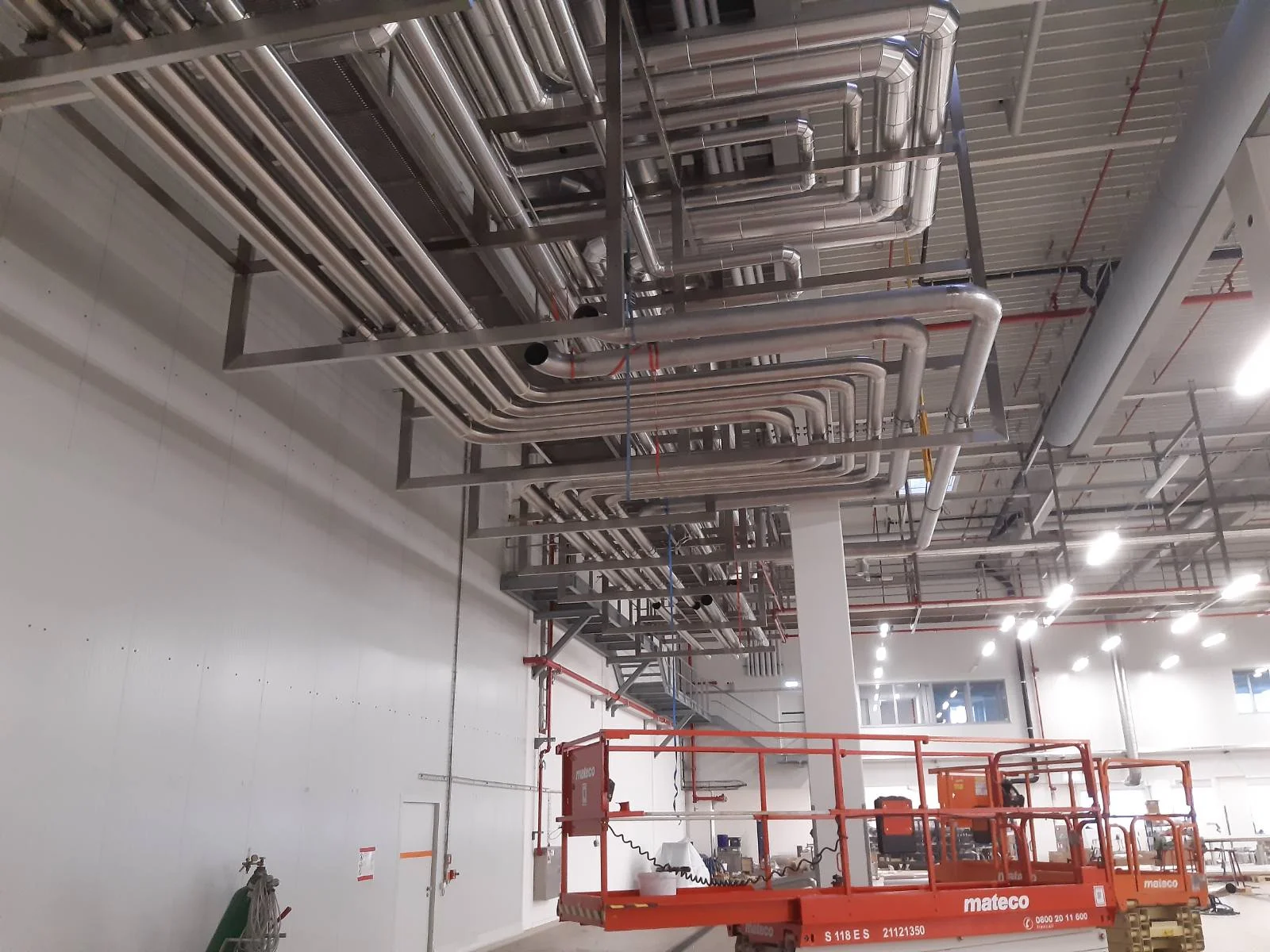 Przemysłowa hala z metalowymi rurami zamontowanymi pod sufitem i pomarańczową platformą hydrauliczną do prac na wysokości.