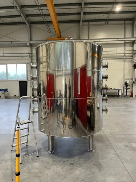 Duży polerowany stalowy zbiornik przemysłowy stojący na trzech podporach w jasnym magazynie z metalową drabiną obok.