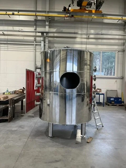 Błyszczący stalowy zbiornik przemysłowy z okrągłym otworem, ustawiony w hali produkcyjnej z regałami i oknem w tle.