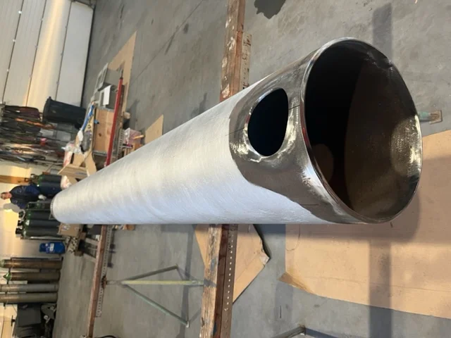 Duży metalowy cylinder ze stali nierdzewnej z okrągłym otworem na środku, stojący w warsztacie przemysłowym.