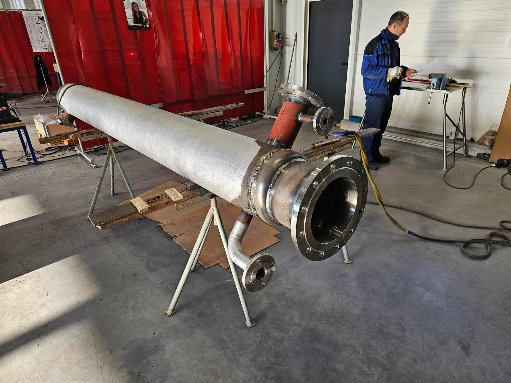 Metalowa rura przemysłowa z kołnierzami na stojakach, mężczyzna w roboczym ubraniu czytający dokumenty w warsztacie.