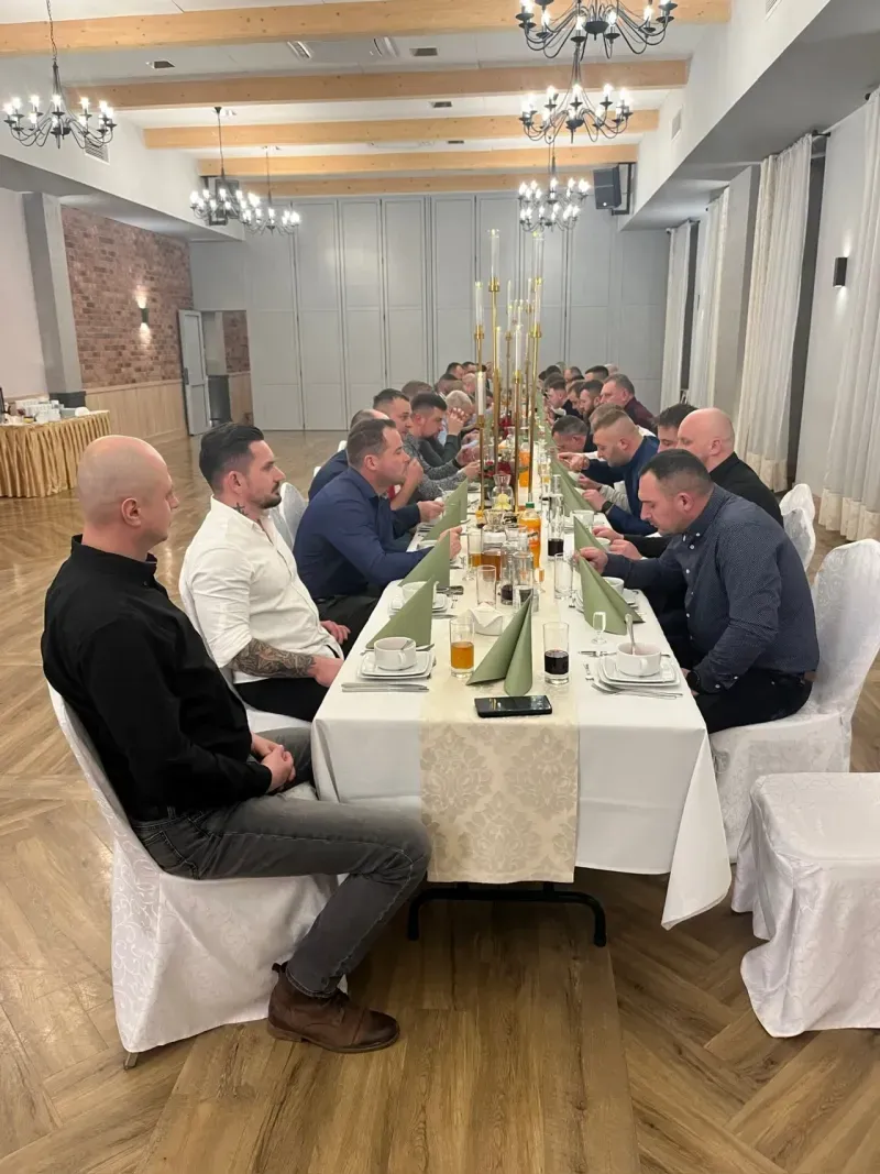 Grupa mężczyzn siedzących po obu stronach długiego stołu w eleganckiej sali, nakrytego białym obrusem i ozdobionego zielonymi serwetkami oraz wysokimi świecznikami.