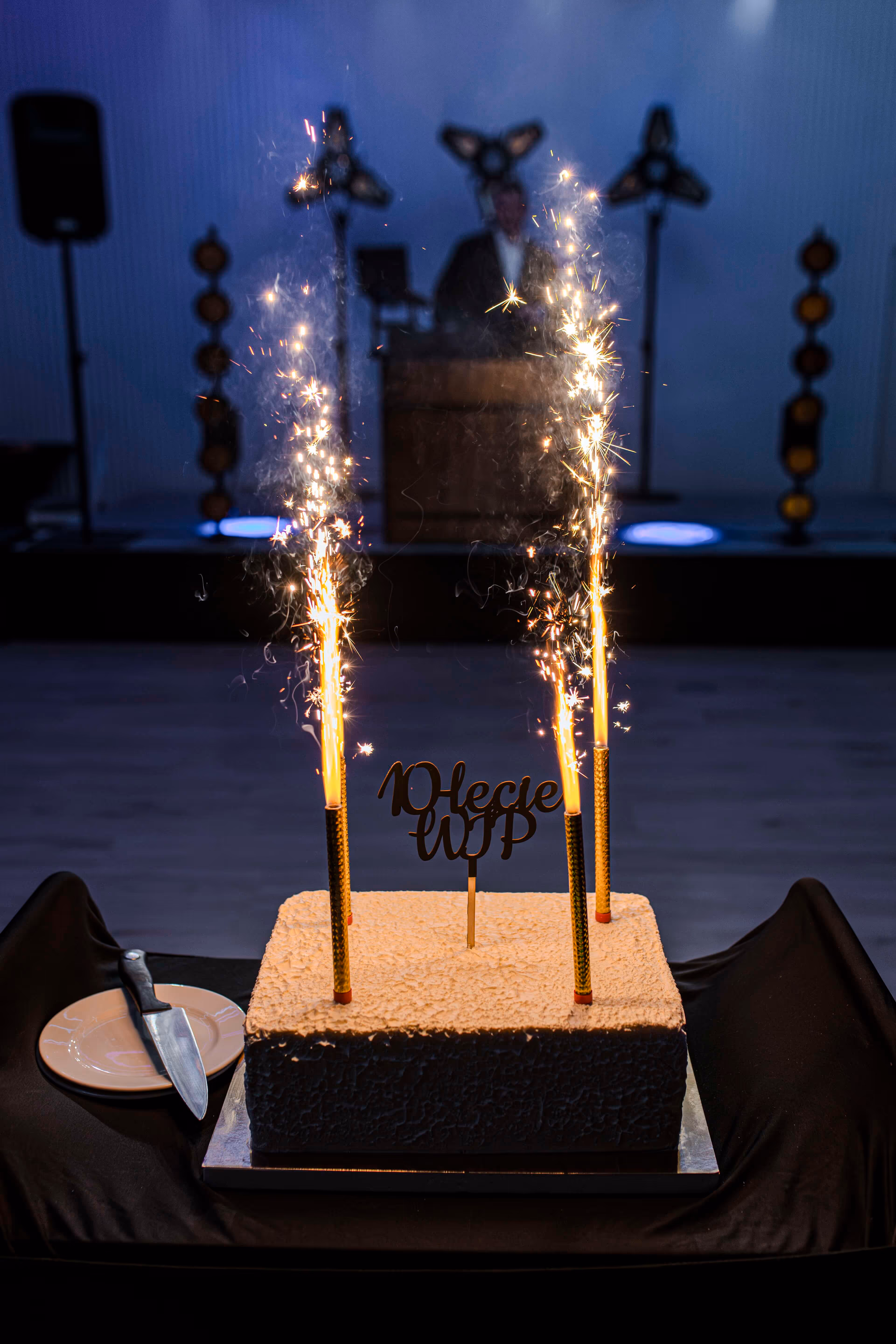 Kwadratowy tort z trzema zapalonymi zimnymi ogniami i ozdobą z napisem „10-lecie WPP”.