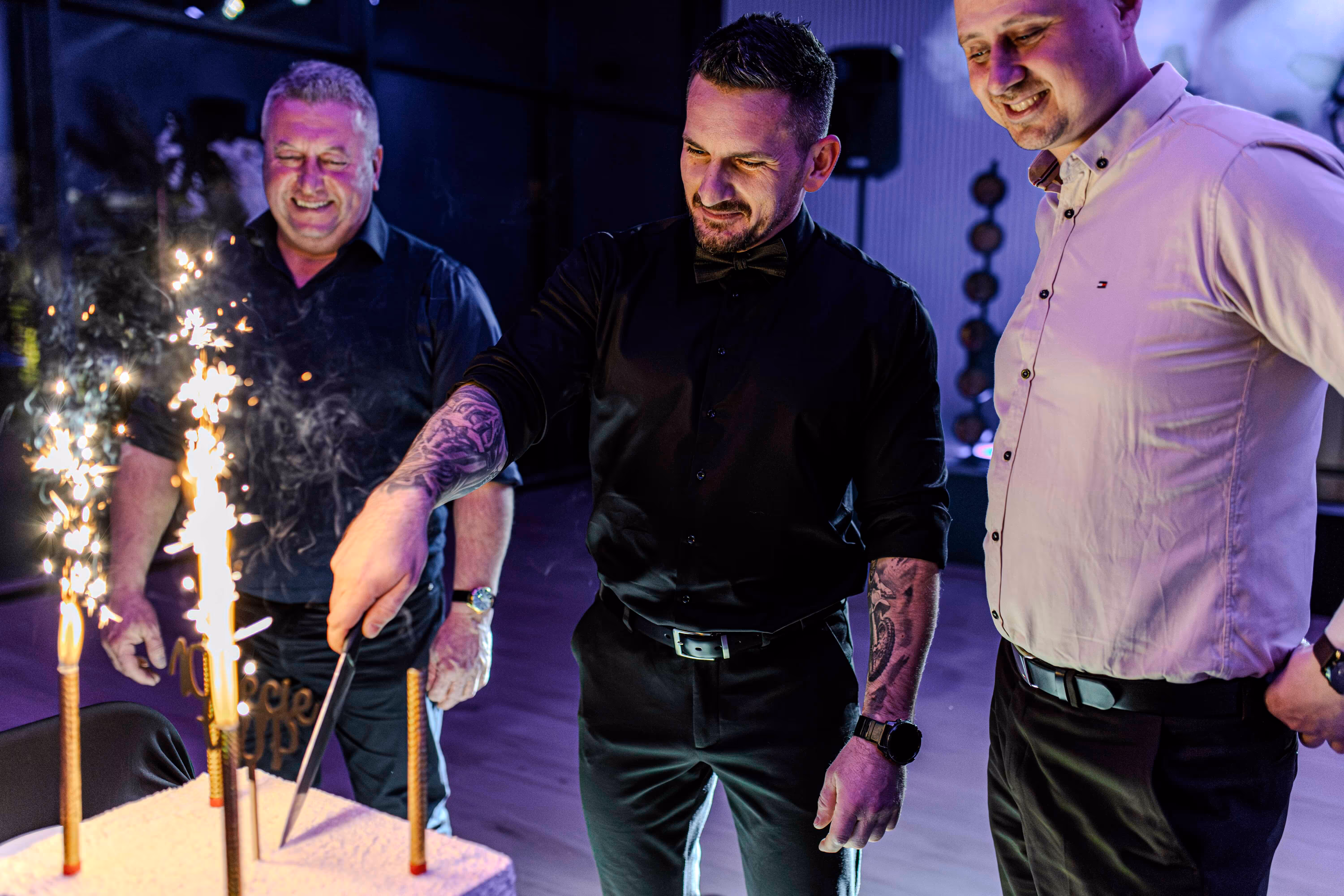 Trzech mężczyzn w eleganckich ubraniach tnie tort z zapalonymi zimnymi ogniami podczas imprezy.