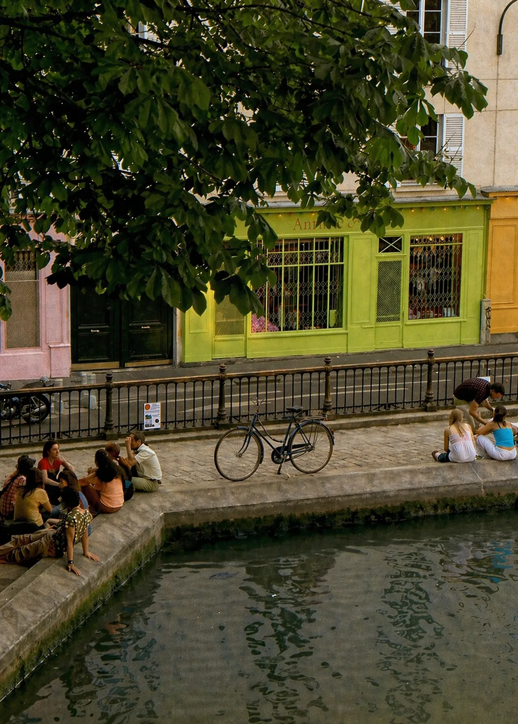 Descente pavée le long du canal Saint-Martin à Paris avec des passants assis au bord de l'eau devant une façade de boutique typique vert amande, capturant l'atmosphère authentique du quartier avec Anchor.