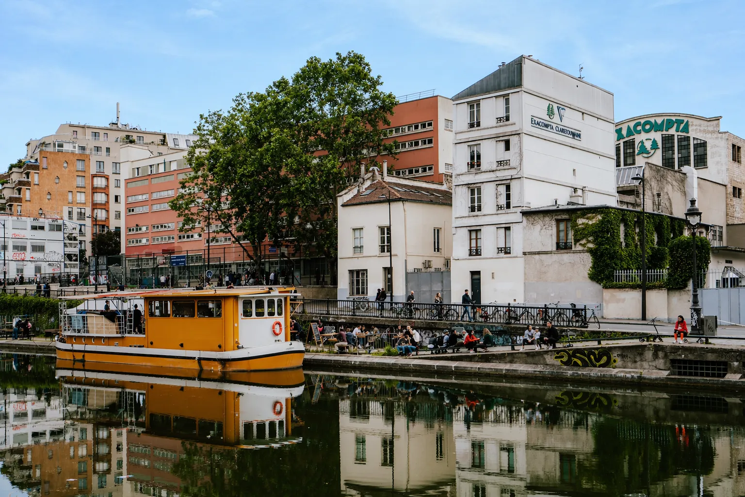 Un bateau traditionnel jaune amarré sur les eaux calmes du canal Saint-Martin à Paris, reflétant l'architecture des quais et les bâtiments historiques environnants comme Exacompta Clairefontaine, une immersion urbaine signée Anchor.