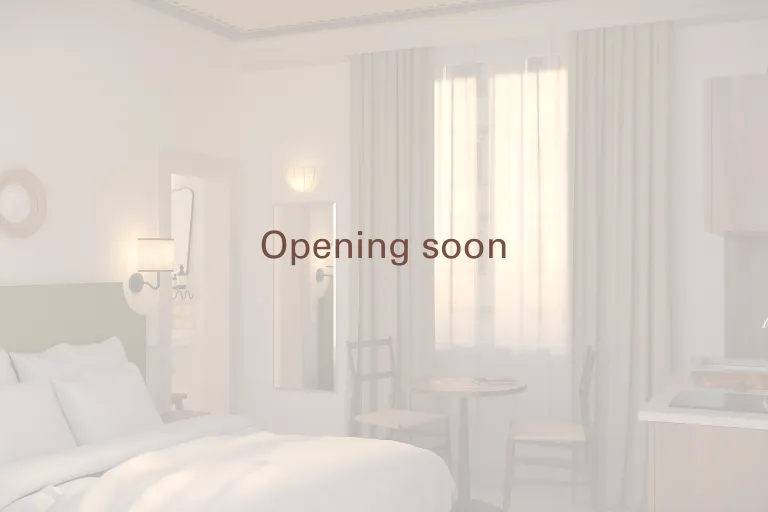 Aperçu en transparence d'un studio haut de gamme Anchor, mettant en valeur un lit double aux draps blancs au premier plan, un miroir mural et un coin repas avec deux chaises près d'une grande fenêtre lumineuse. L'inscription « Opening soon » est centrée su