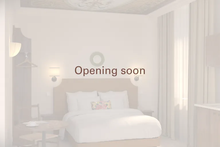 Aperçu en transparence d'une chambre élégante et lumineuse d'un hébergement haut de gamme Anchor, avec l'inscription au centre « Opening soon » surmontée d'un logo circulaire épuré. La pièce comprend un lit double avec une tête de lit beige, des oreillers
