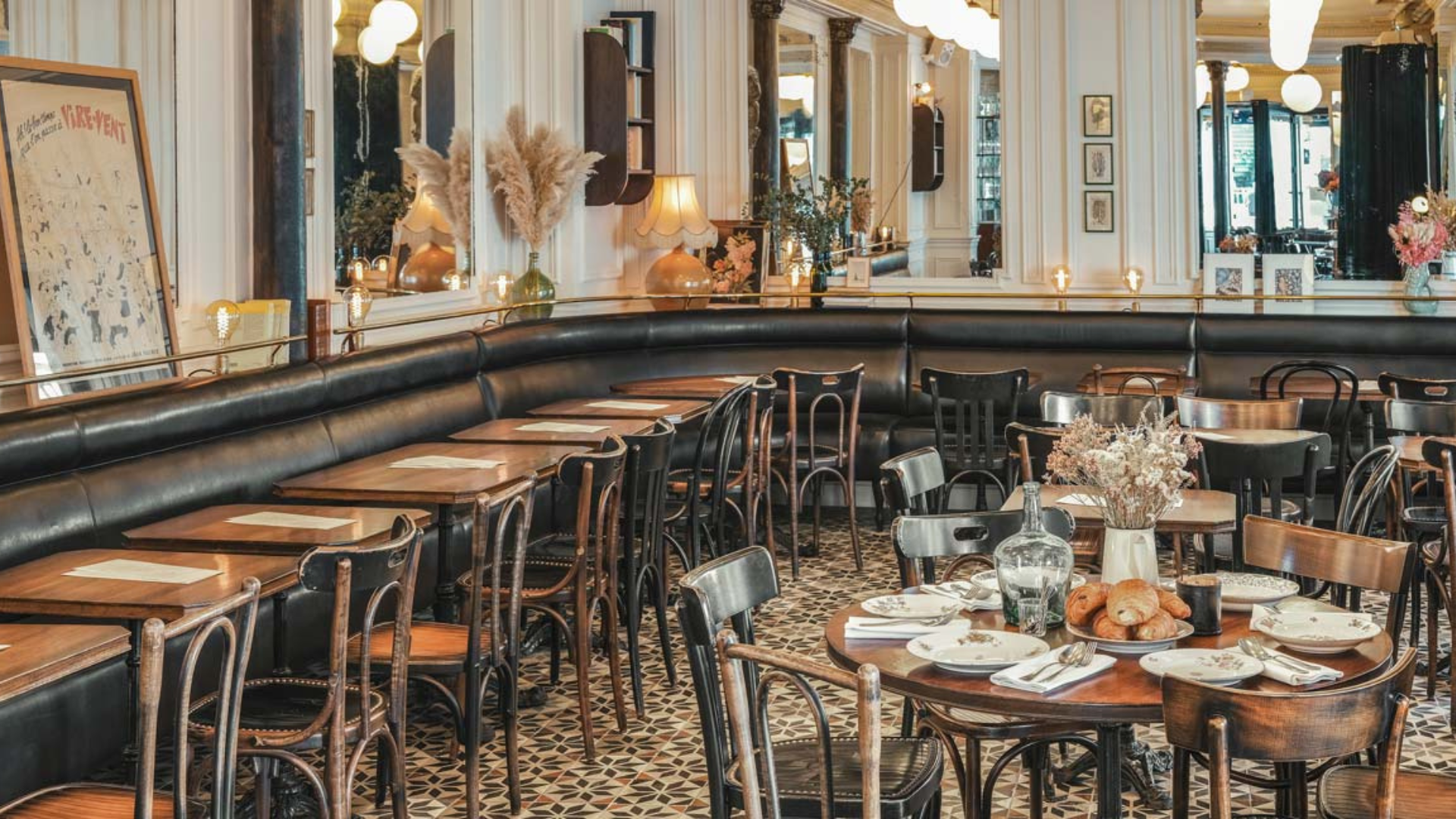 Salle d'un brasserie parisienne rétro avec banquettes en cuir noir, chaises bistrot en bois courbé, tables en bois, sol en carreaux de ciment géométriques, grands miroirs et suspensions boules blanches