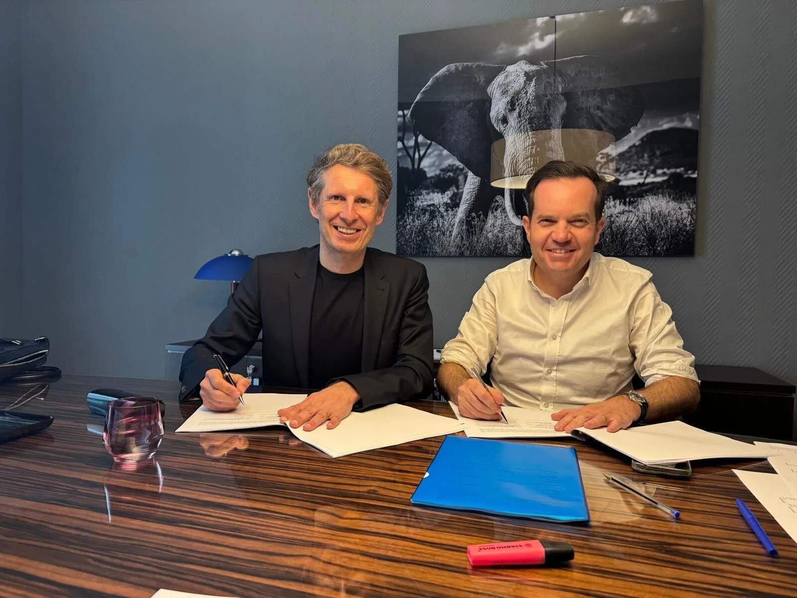 Two men sitting at a polished wooden table, signing documents and smiling, with a black and white elephant photo on the wall behind them.