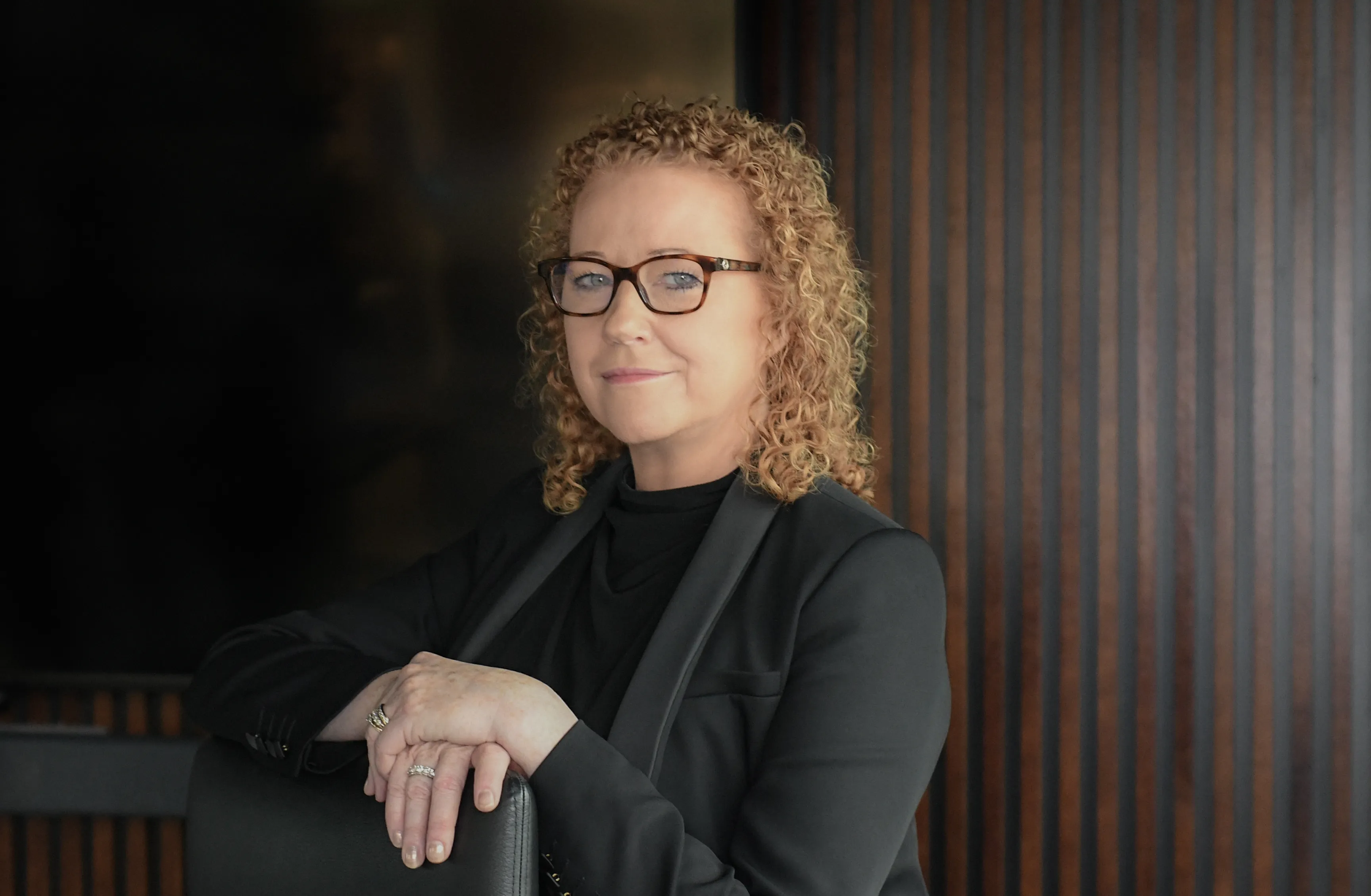 Woman with curly hair and glasses wearing a black blazer, sitting with hands resting on a chair back.