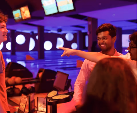 Group of friends smiling and pointing while enjoying a bowling alley with neon lighting.