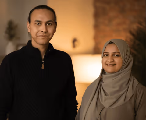 A man with a dark zip-up sweater and a woman wearing a beige hijab stand side by side in a warmly lit room.