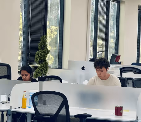 Two people working on Apple iMac computers in a bright office with large windows and modern office chairs.
