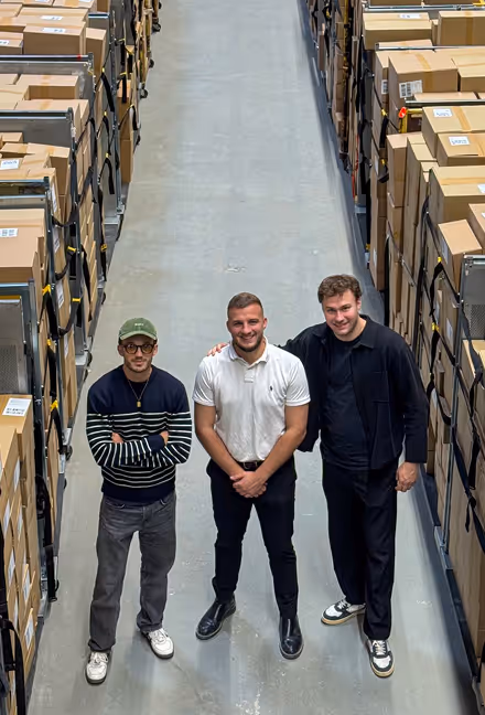 Three men standing smiling together in a warehouse aisle filled with stacked cardboard boxes.