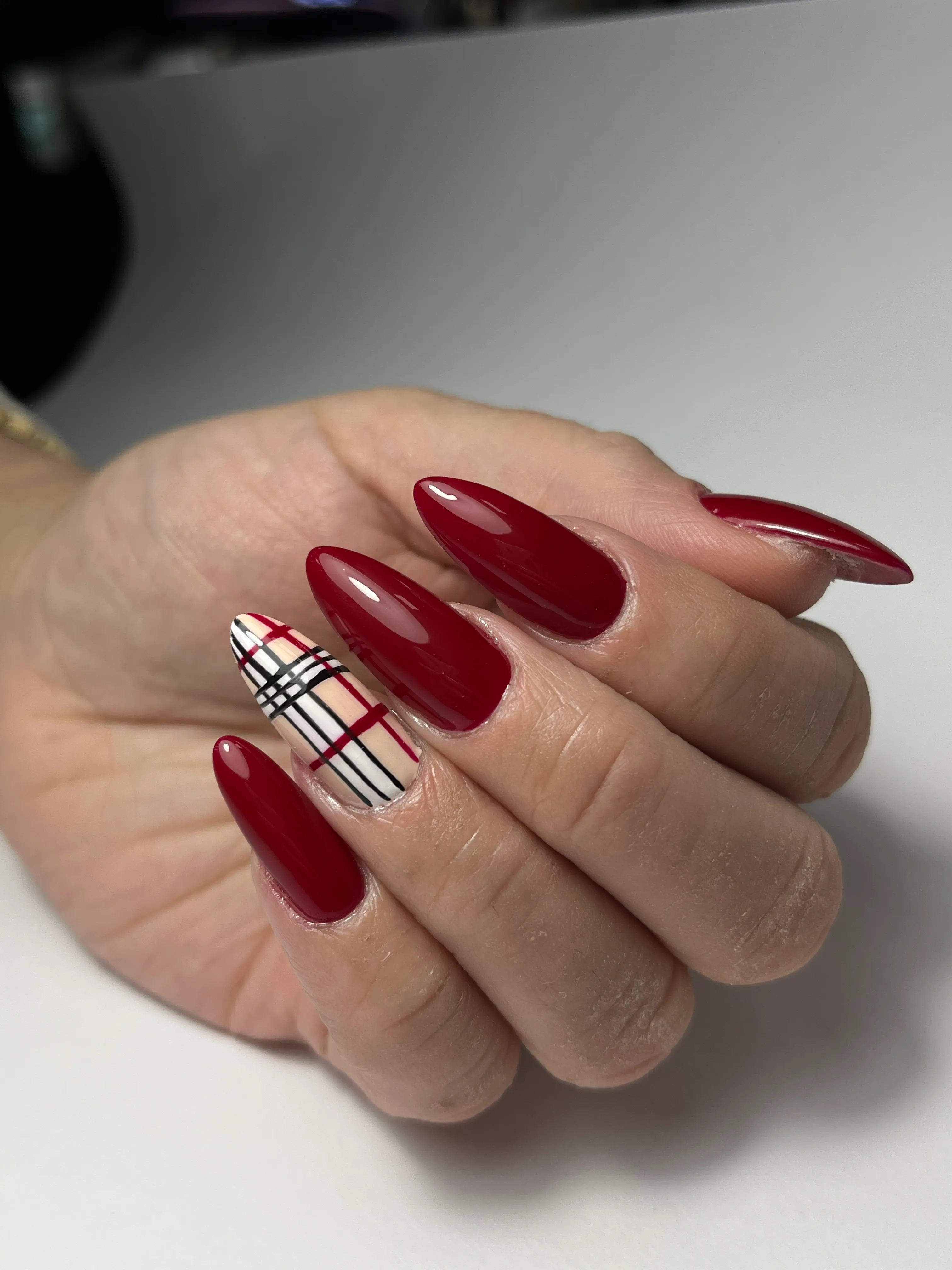 Hand with long almond-shaped nails painted glossy dark red, with one nail featuring a beige base and red and black plaid lines.