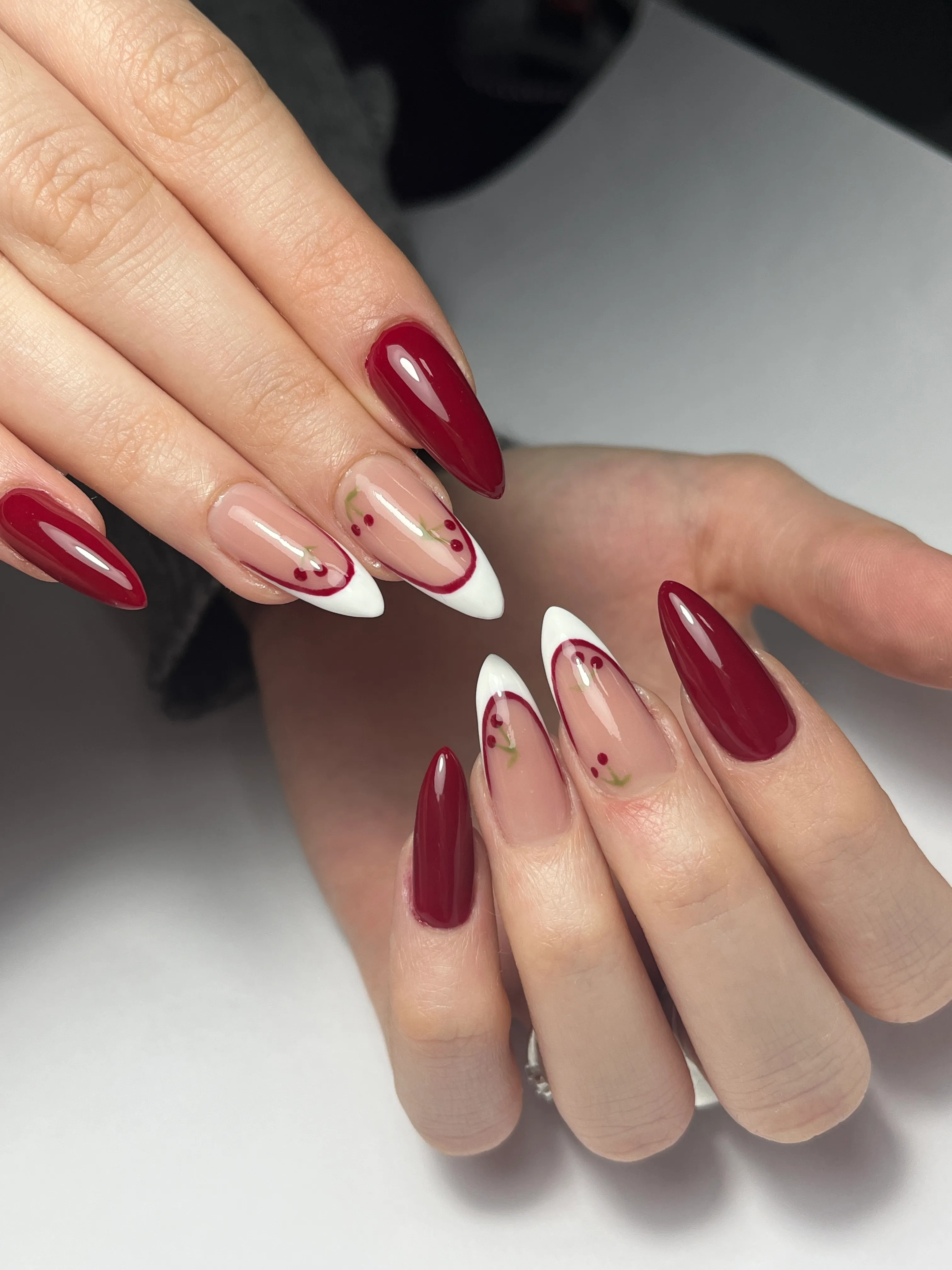 Hands with long almond-shaped nails painted deep red, with some nails featuring white tips and delicate red berry designs on a nude base.