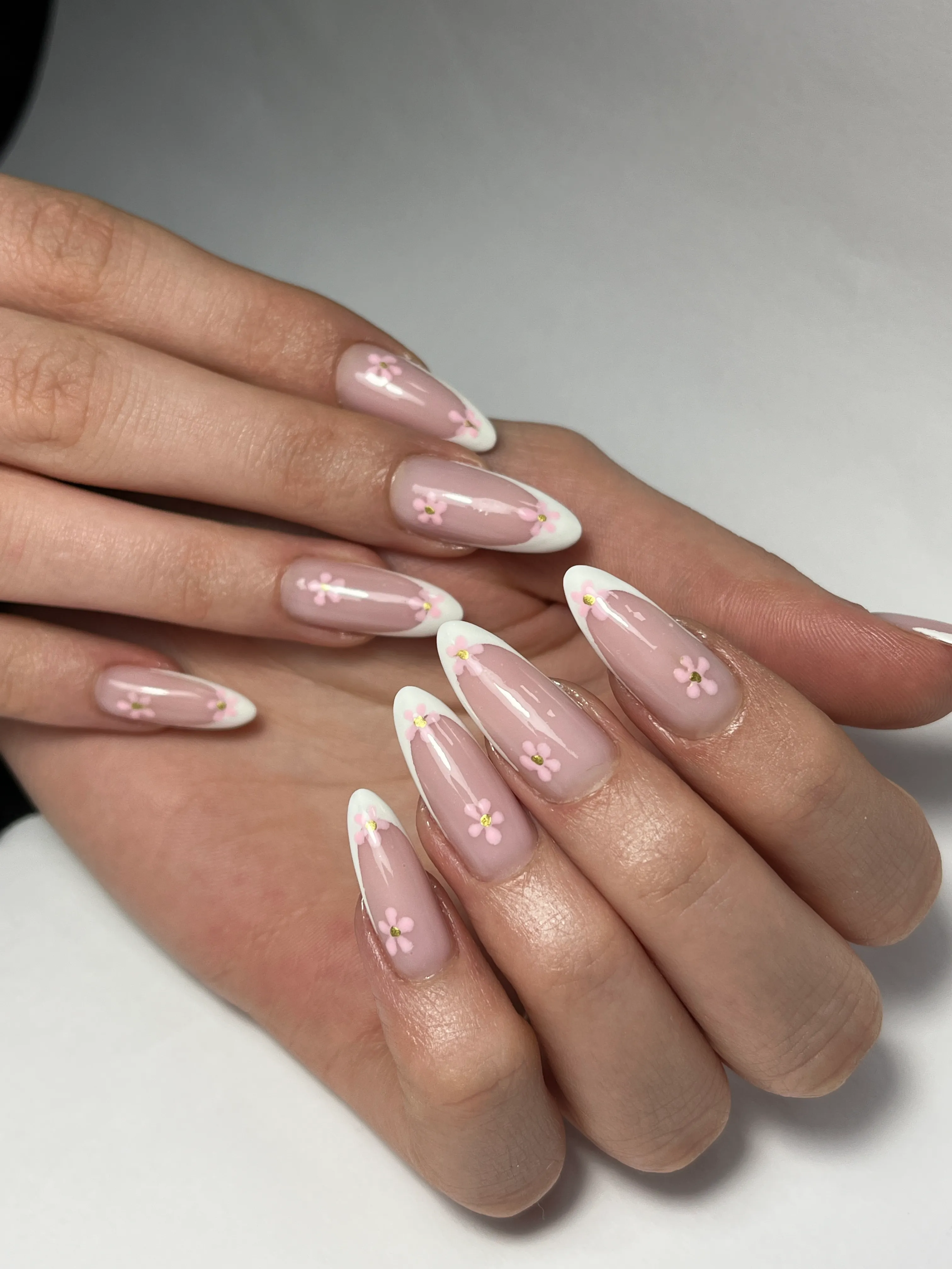 Hands with almond-shaped nails featuring a pink French tip manicure decorated with small pink flower designs and gold centers.