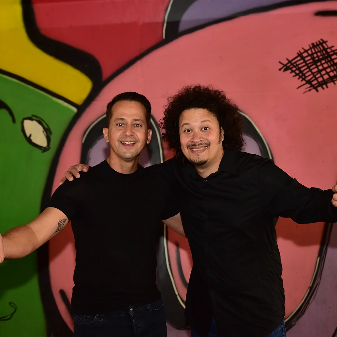 Two men smiling with arms around each other in front of a colorful graffiti wall.