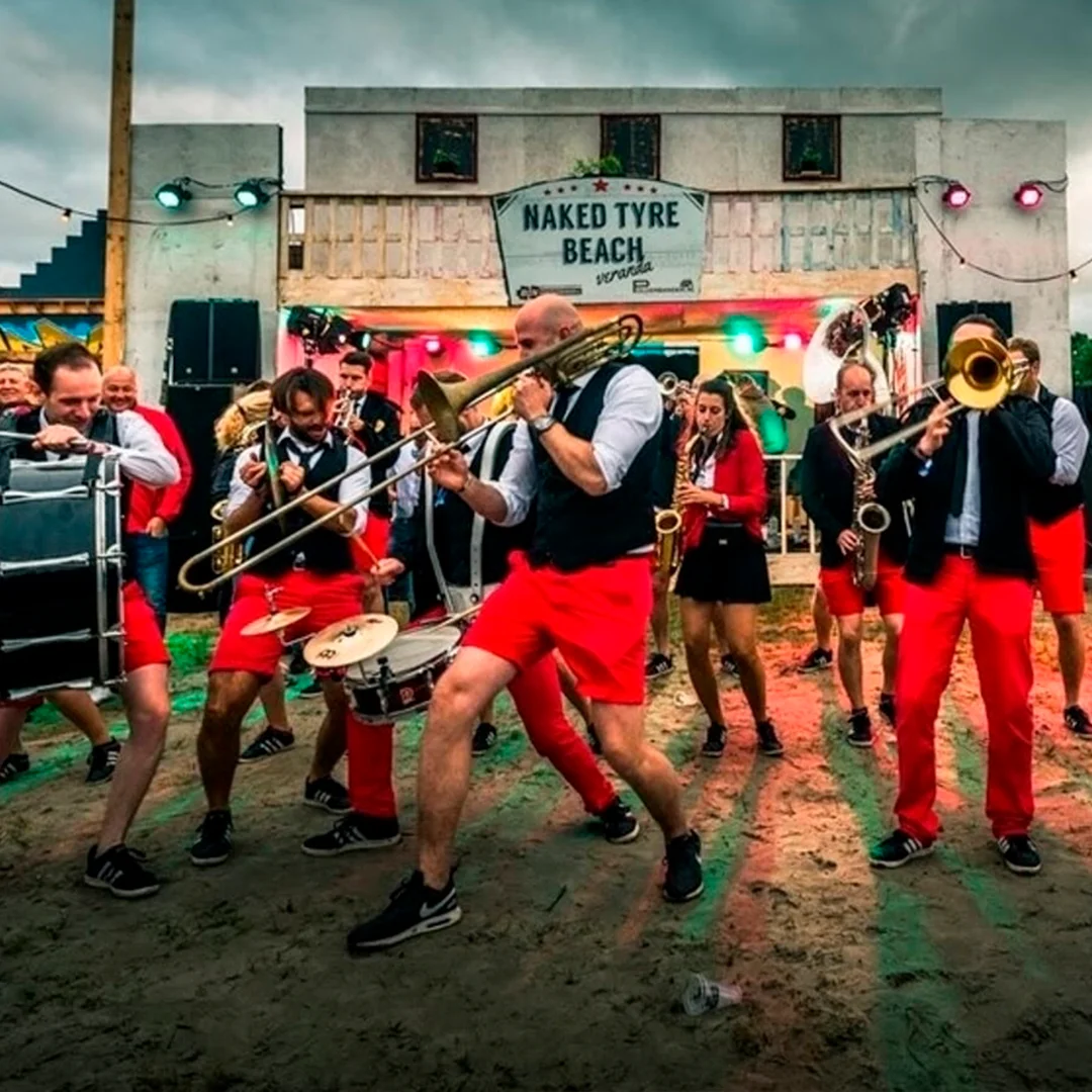 Band with brass instruments and drums wearing red shorts playing energetically outdoors on sandy ground.