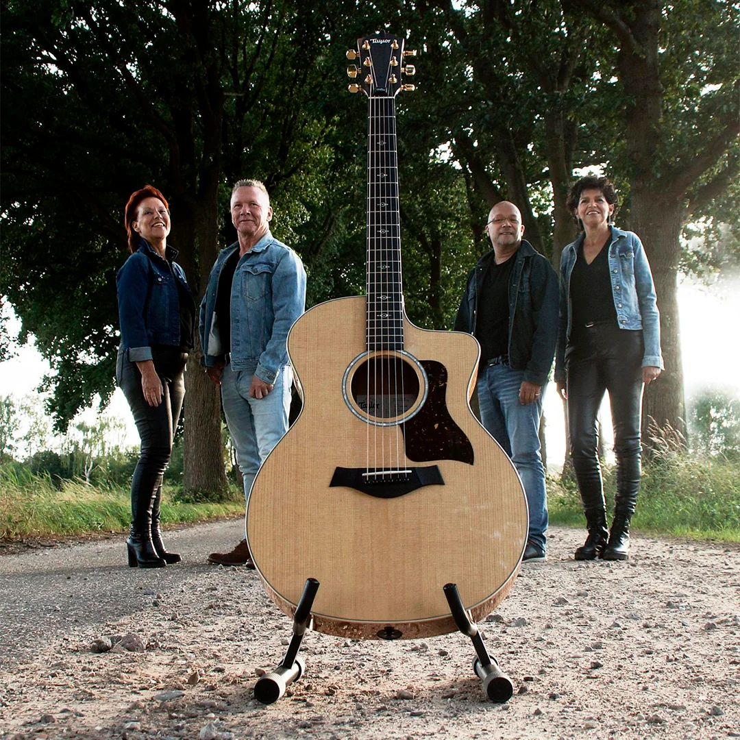 Four adults standing behind an acoustic guitar on a stand in a tree-lined outdoor setting.