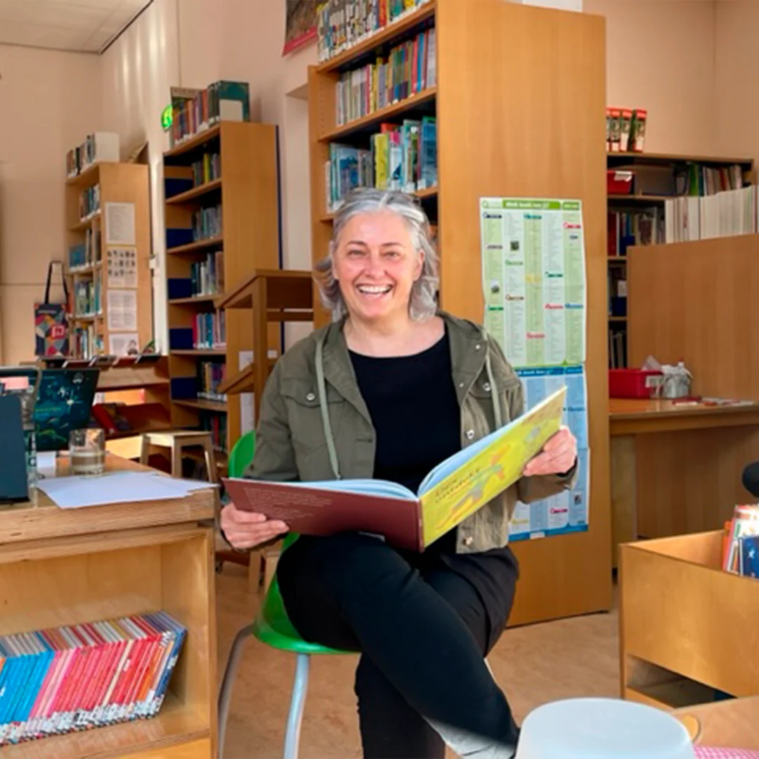 Smiling woman reading a colorful book in a library with shelves of books behind her.