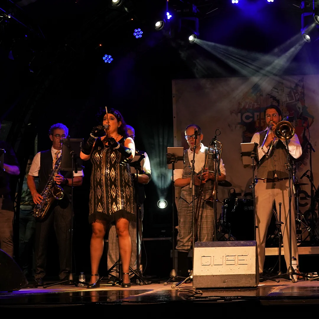 Band performing on stage with female singer and musicians playing saxophone and trombone under spotlights.