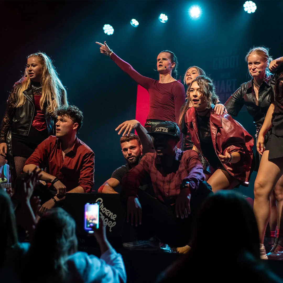 Group of young performers on stage under bright lights with audience in foreground.