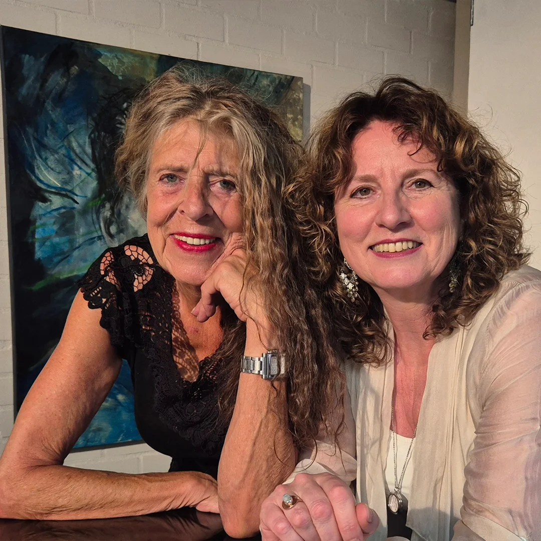 Two smiling women with curly hair leaning on a table, one in black lace and the other in light clothing.