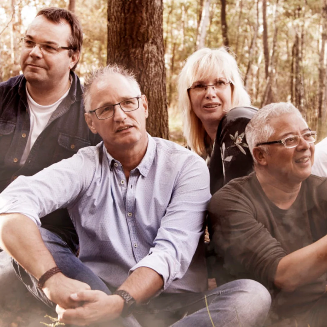 Four adults sitting and smiling outdoors among trees in a relaxed group setting.