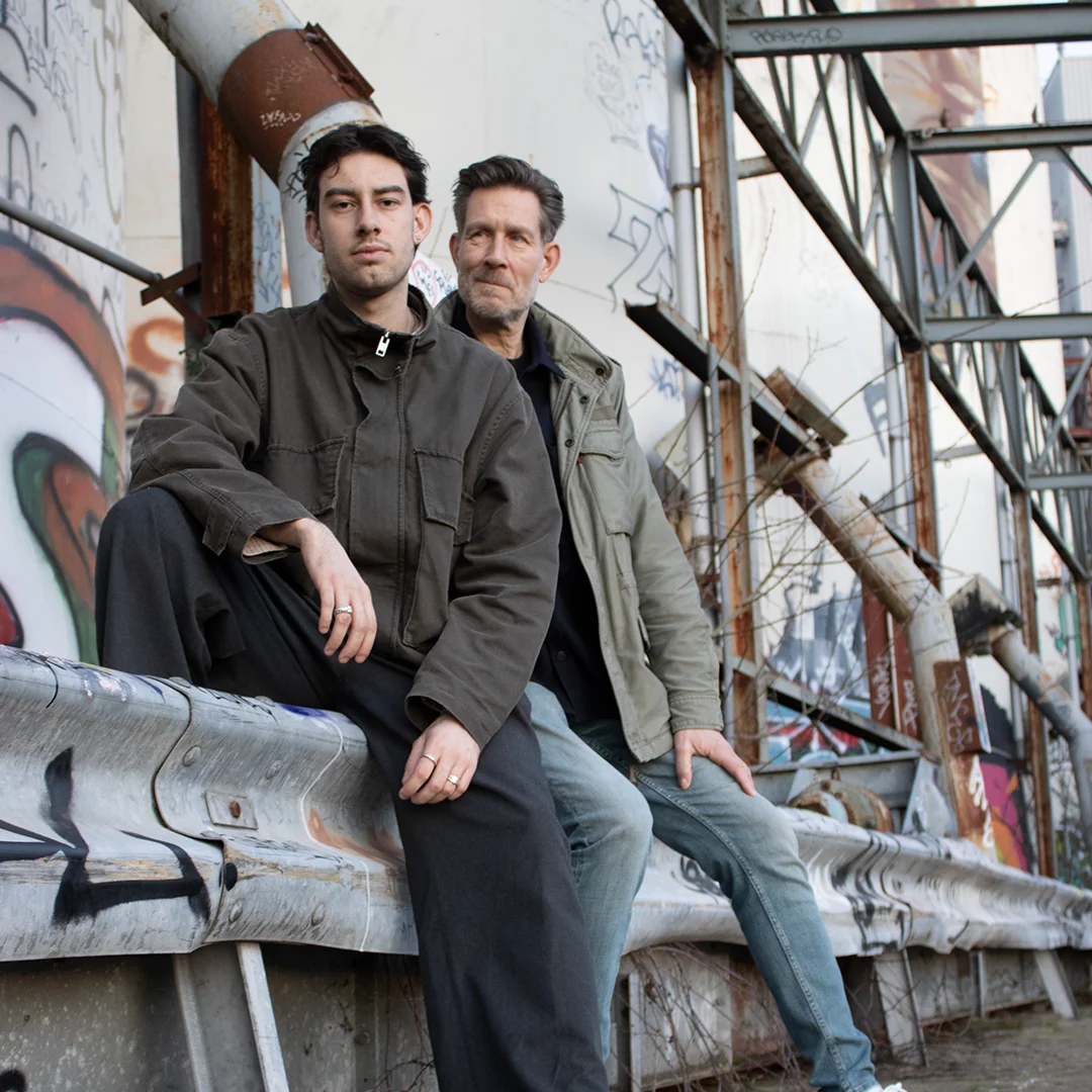 Two men in casual jackets sitting on a graffiti-covered metal guardrail in an urban area.