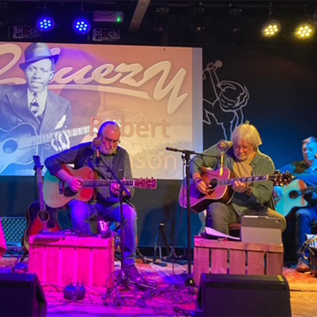 Three men seated on stage playing acoustic guitars under stage lights in front of a blues-themed backdrop.