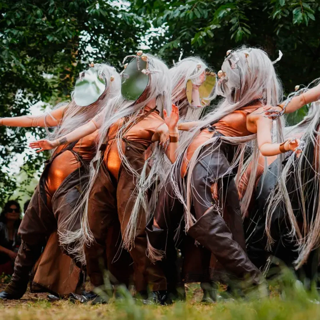 Performers with long white hair and reflective masks dance in brown costumes outdoors among trees.